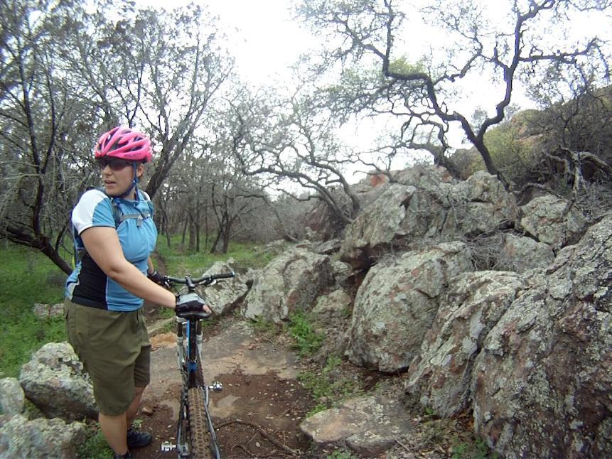 A person wearing a pink helmet and a blue cycling jersey stands beside a mountain bike on a narrow trail surrounded by rocks and trees. They appear to be looking to the side, with a green grassy area visible in the background. The scene is set in a natural outdoor environment, likely for mountain biking. Reveille Peak Ranch mountain bike trail.