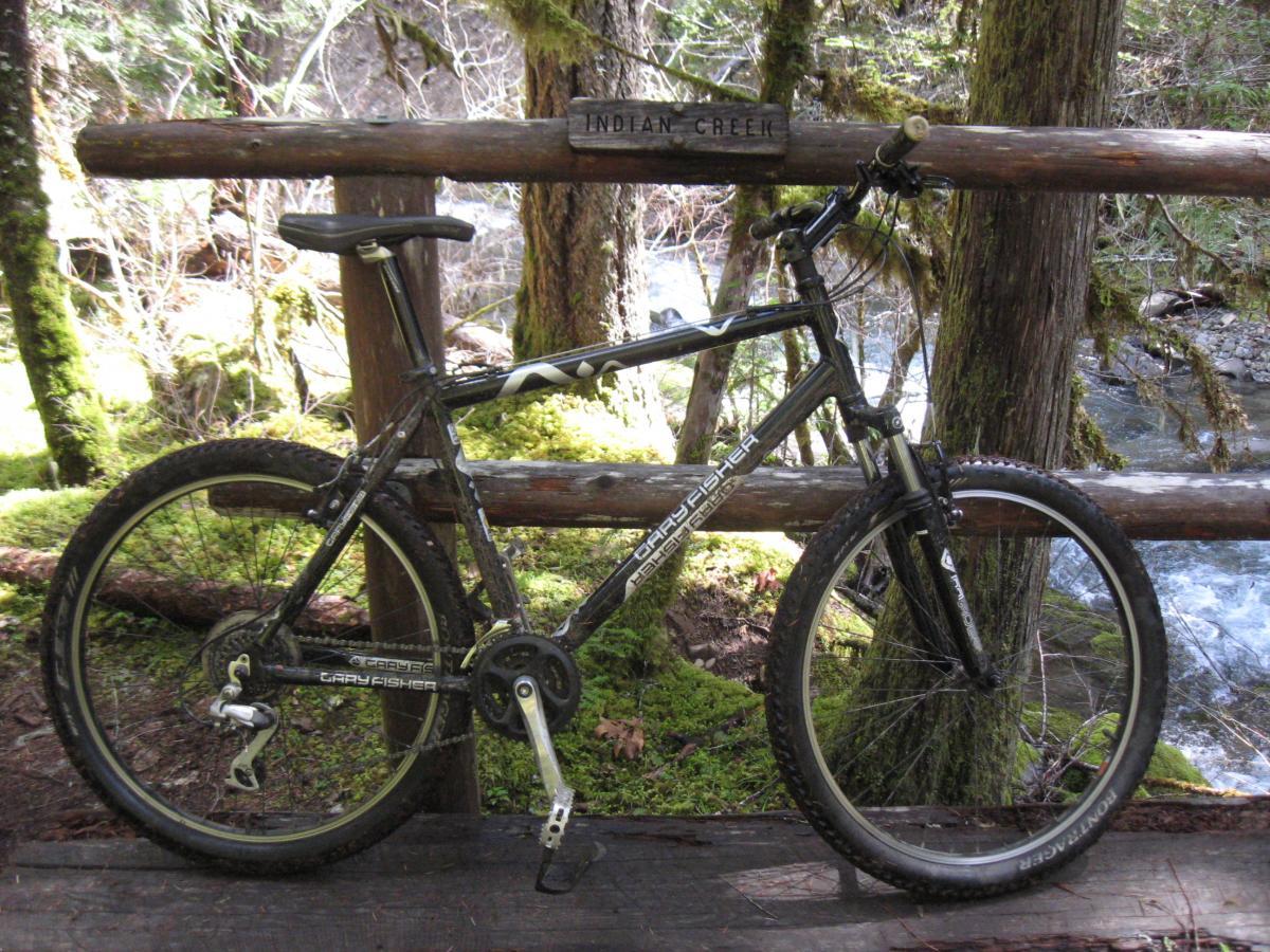 A mountain bike leaning against a wooden railing labeled "Indian Creek," surrounded by lush green foliage and trees, with a stream visible in the background. Middle Fork Trail mountain bike trail.