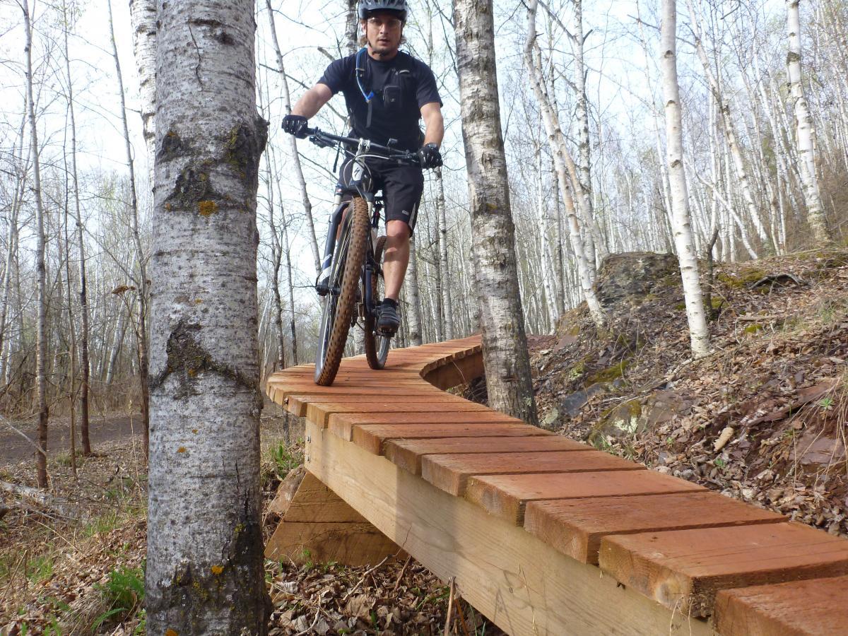 A mountain biker rides on a wooden bridge through a forest, surrounded by birch trees. The cyclist is wearing a helmet and sports gear, showcasing skill as they navigate the elevated path in a natural setting. Cuyuna Lakes mountain bike trail.