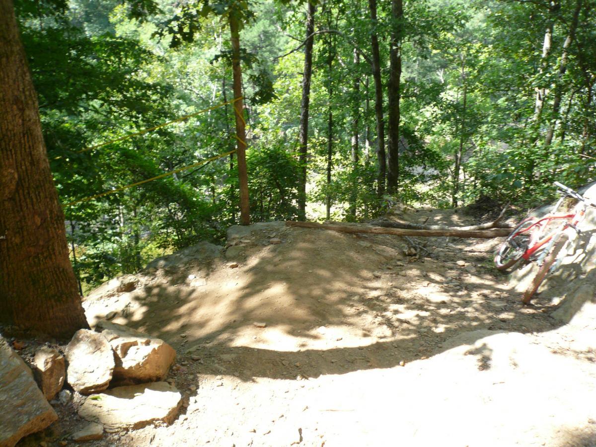 A dirt bike trail surrounded by trees, featuring a small jump and a red bicycle leaning against a log. Sunlight filters through the foliage, casting shadows on the path. The scene conveys a sense of adventure and outdoor activity in a forested area. Van Michael Trail mountain bike trail.