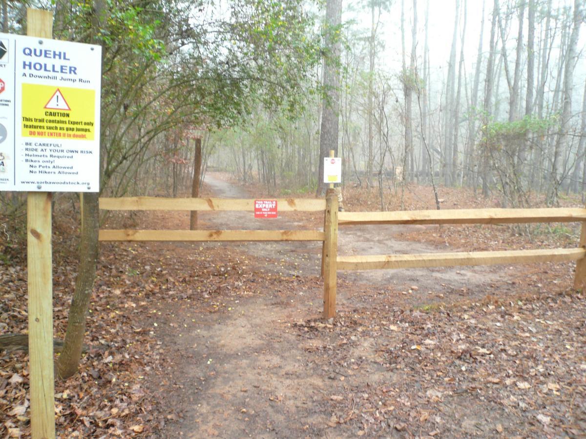 A wooden entrance gate leading to a wooded trail marked as "QUEHL HOLLER," a downhill jump run. A caution sign indicates the trail is for expert riders only, with warnings about terrain features like gap jumps and advises riders to enter at their own risk. The surrounding area is misty and forested, with fallen leaves on the ground. Quehl Holler mountain bike trail.