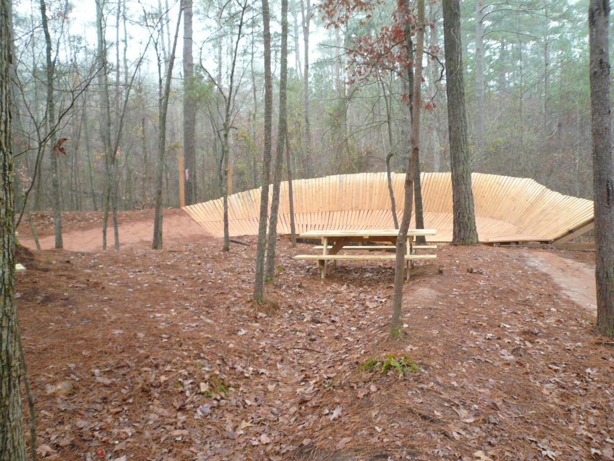 A wooden bike ramp set in a foggy forest, surrounded by tall trees and a layer of pine needles on the ground. A picnic table is positioned nearby, offering a place to rest. Quehl Holler mountain bike trail.
