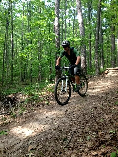 A person riding a mountain bike on a dirt trail in a wooded area, surrounded by green trees and foliage. The cyclist is wearing a helmet and athletic clothing. Blankets Creek mountain bike trail.