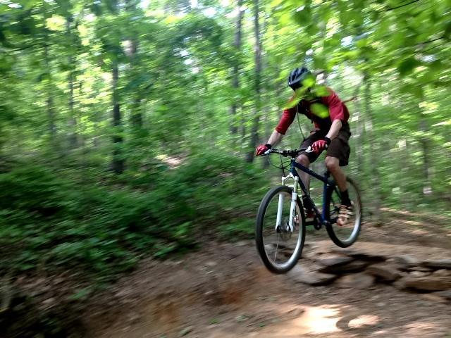 A mountain biker wearing a helmet and protective gear is airborne while jumping off a rocky section of a dirt trail in a lush green forest. Sunlight filters through the tree canopy, illuminating the scene. Blankets Creek mountain bike trail.
