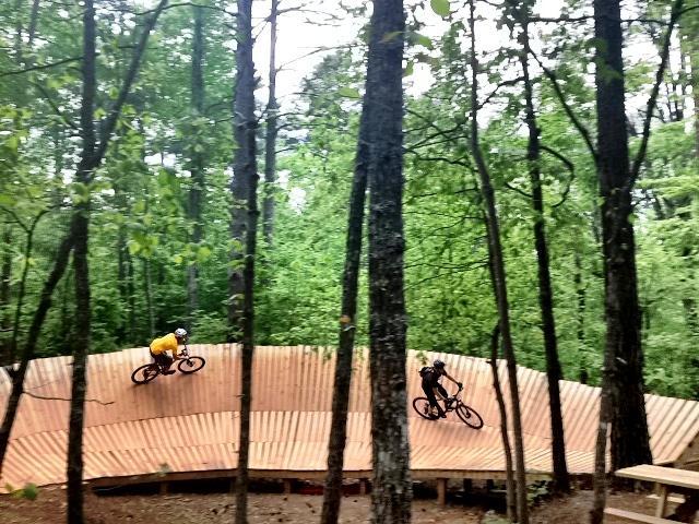 Two mountain bikers riding on a wooden pump track situated in a lush green forest. One rider is wearing a yellow shirt, while the other is in darker clothing. The track features a series of curved wooden sections that allow for dynamic riding, surrounded by tall trees and foliage. Blankets Creek mountain bike trail.