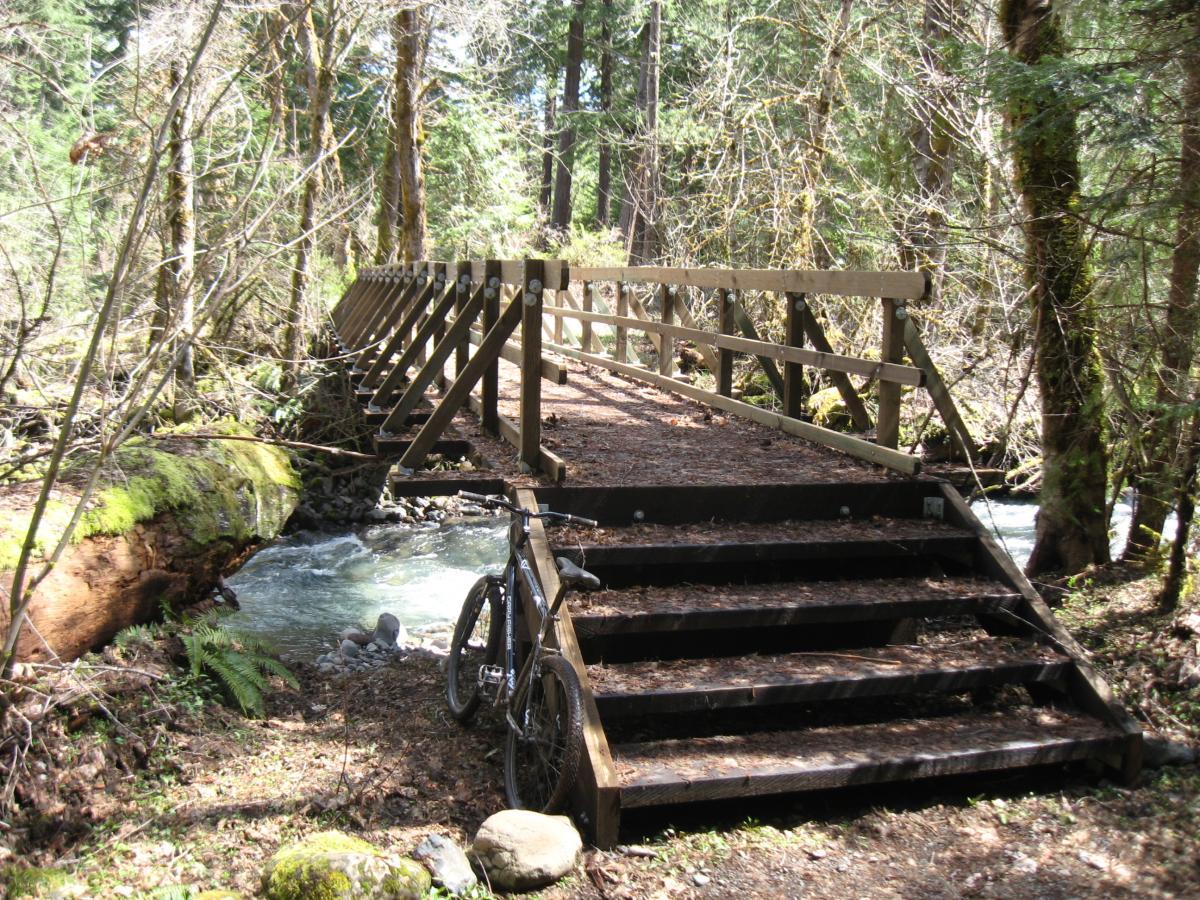 A wooden bridge over a small creek surrounded by trees and foliage, with a bike resting on the ground near the steps leading up to the bridge. Middle Fork Trail mountain bike trail.