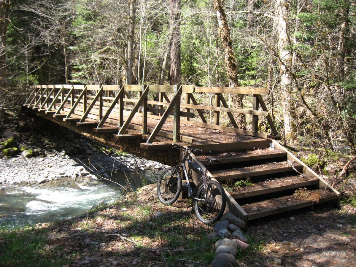 A wooden bridge spans a small creek in a forested area, with lush greenery and trees surrounding the scene. A bicycle is leaned against the steps of the bridge, and smooth stones line the water's edge. Sunlight filters through the trees, creating a peaceful outdoor atmosphere. Middle Fork Trail mountain bike trail.