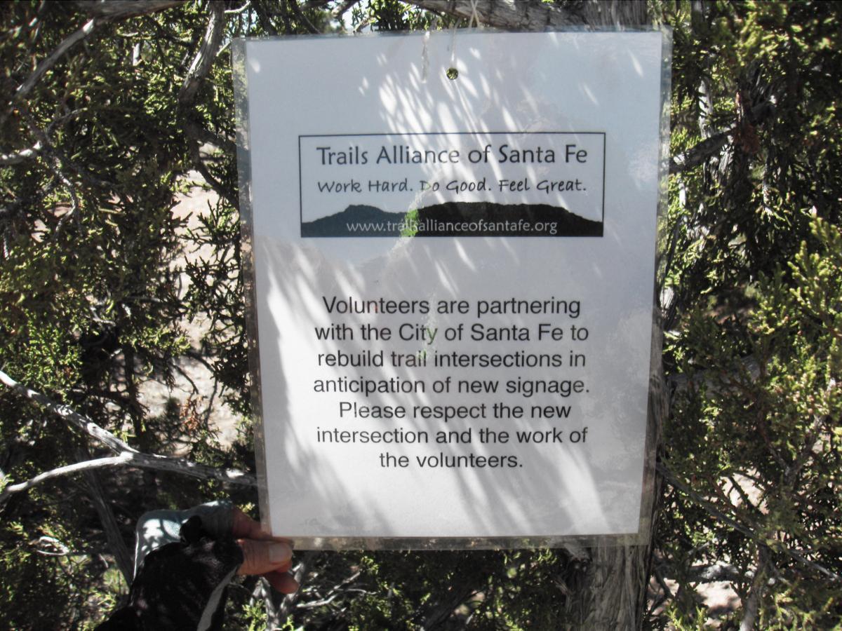 Sign posted in a natural area, promoting the Trails Alliance of Santa Fe. It informs visitors that volunteers are collaborating with the City of Santa Fe to rebuild trail intersections for new signage and requests respect for the volunteers' work. La Tierra mountain bike trail.