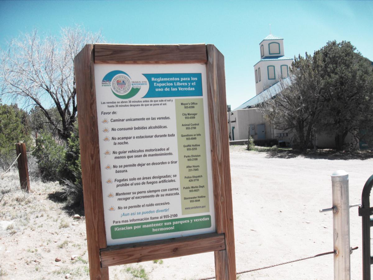 Sign displaying regulations for the use of trails and open spaces, written in Spanish. It includes guidelines such as walking only on paths, not consuming alcohol, camping restrictions, and maintaining noise levels. A building and trees are visible in the background, indicating a park or recreational area. La Tierra mountain bike trail.