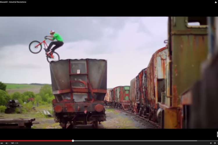 A mountain biker performing a jump over a train wagon in an industrial setting, with a backdrop of cloudy skies and abandoned train cars.