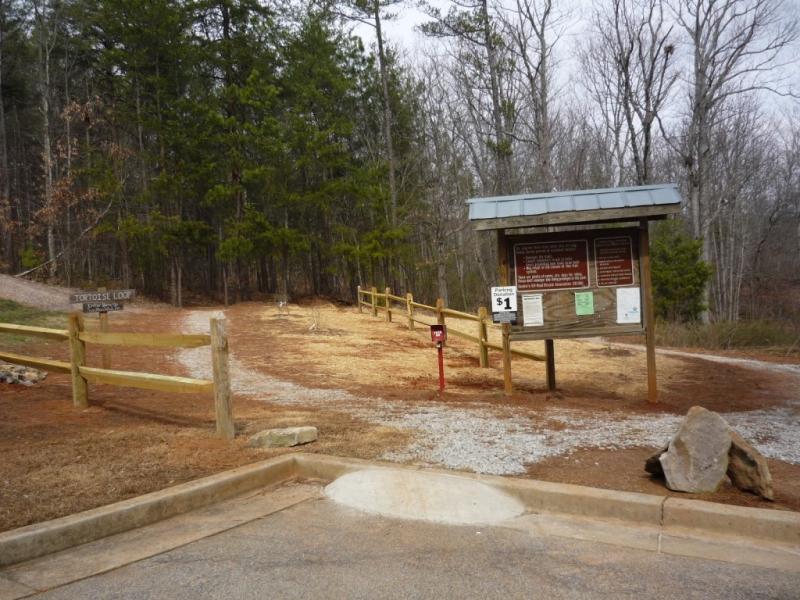 Entrance to a hiking trail marked with a sign displaying information and rules. A wooden fence lines the path, which leads into a wooded area. There are gravel and dirt surfaces, and a small fee collection box is visible. Surrounding trees are sparse, indicating a transitional season.