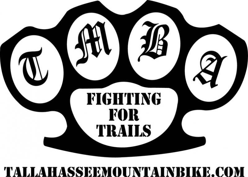 Logo featuring a stylized set of brass knuckles with the letters "TMBA" inside the round sections, accompanied by the text "FIGHTING FOR TRAILS" prominently displayed in the center. Below, the website "tallahasseemountainbike.com" is included. The design is bold and monochromatic, emphasizing a strong connection to mountain biking and trail advocacy.