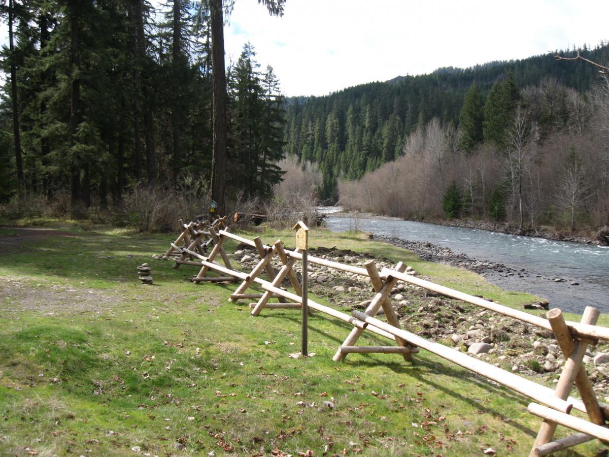 A serene riverside scene featuring a wooden fence along a grassy area, with scattered rocks and trees in the background. The river flows gently, surrounded by a mix of evergreen and deciduous trees, under a partly cloudy sky. Middle Fork Trail mountain bike trail.