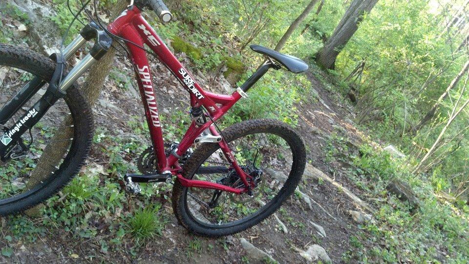 A red mountain bike is parked on a dirt trail surrounded by green foliage and trees. The bike features front suspension and is positioned near a rocky area, suggesting it is meant for off-road riding. Sugar Hollow Park mountain bike trail.