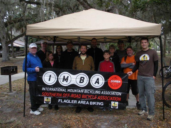 A group of people pose under a canopy, holding a banner for the International Mountain Bicycling Association (IMBA) and the Southern Off-Road Bicycle Association (SORBA). The background features trees and a park setting, and the group appears to be gathered for a cycling-related event.