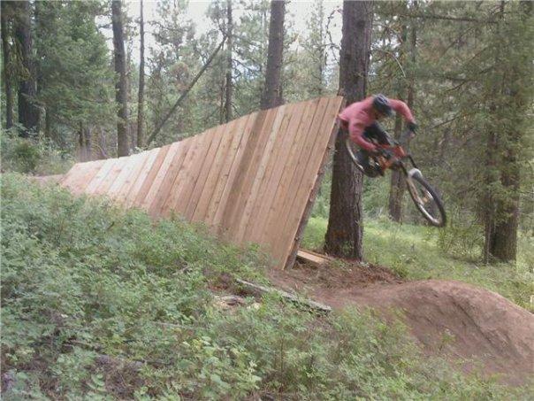 A mountain biker performs a jump off a wooden ramp in a forested area. The bike is airborne as it descends from the ramp, surrounded by trees and greenery.