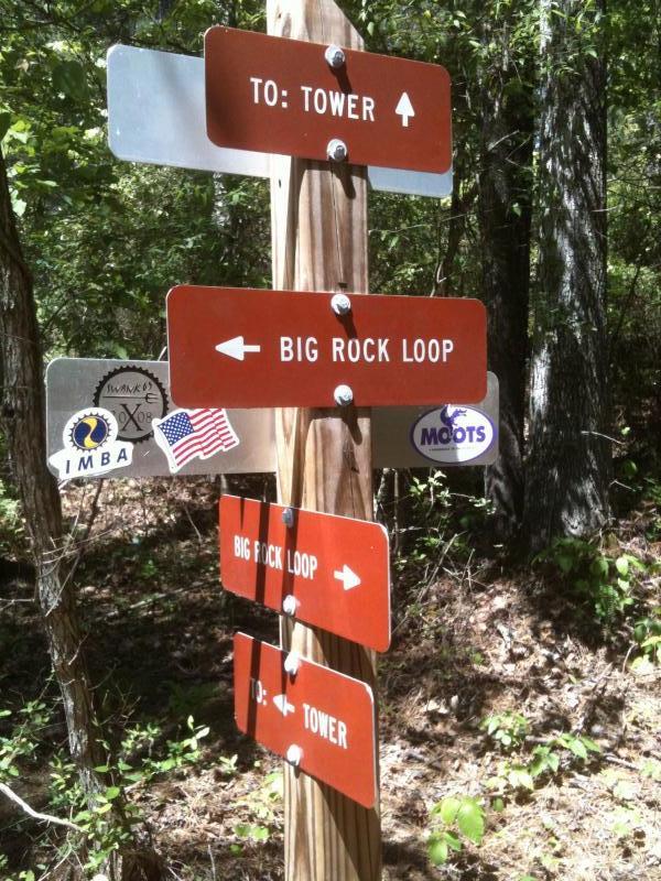 A wooden signpost in a forest with multiple directional arrows. The signs indicate routes to "TOWER" and "BIG ROCK LOOP," with stickers from cycling organizations and brands attached to the post. The surrounding area features trees and natural vegetation. Forks Area Trail System (FATS) mountain bike trail.