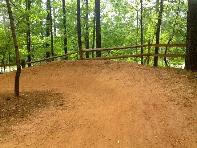 A dirt path winding through a wooded area, featuring a raised, curved mound of dirt, with a wooden fence running alongside it. The background is filled with lush green trees, providing a natural and serene setting. Big Creek mountain bike trail.