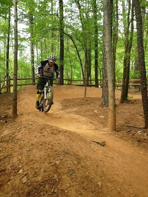 A mountain biker navigating a dirt trail surrounded by trees, jumping off a small ramp. The rider is wearing a helmet and protective gear, and the ground is a reddish-brown color typical of dirt bike paths. The scene is set in a green, wooded area, showcasing a vibrant natural environment. Big Creek mountain bike trail.