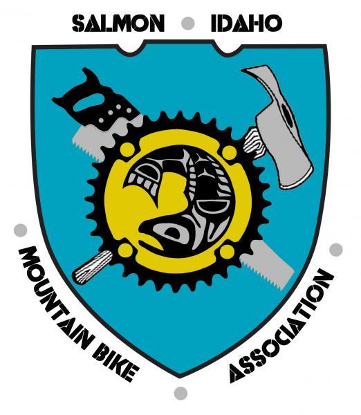 Logo of the Salmon Idaho Mountain Bike Association, featuring a shield design. The shield is divided into a blue background with a central yellow circle displaying a stylized fish graphic. Surrounding the fish are various tools, including a saw and an axe, representing outdoor activities. The words "Salmon," "Idaho," and "Mountain Bike Association" are prominently displayed above and below the shield.