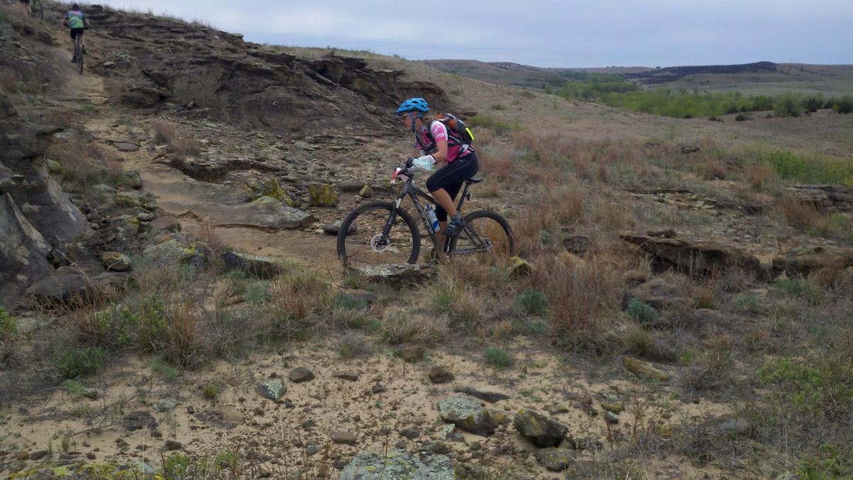 A mountain biker navigating a rocky trail in a grassy landscape, with a second biker visible in the background. The cyclist is wearing a blue helmet and a colorful jersey, riding over uneven terrain with scattered rocks and dry grass. The sky is overcast, suggesting an early morning or late afternoon. Switchgrass mountain bike trail.