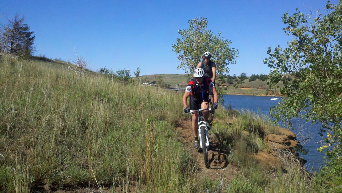 Two mountain bikers navigating a narrow trail along a lakeshore. Lush green grass and a few trees line the path, with a clear blue sky overhead. One biker is in the foreground, focused on the trail, while the other follows closely behind. The water of the lake is visible in the background. Switchgrass mountain bike trail.