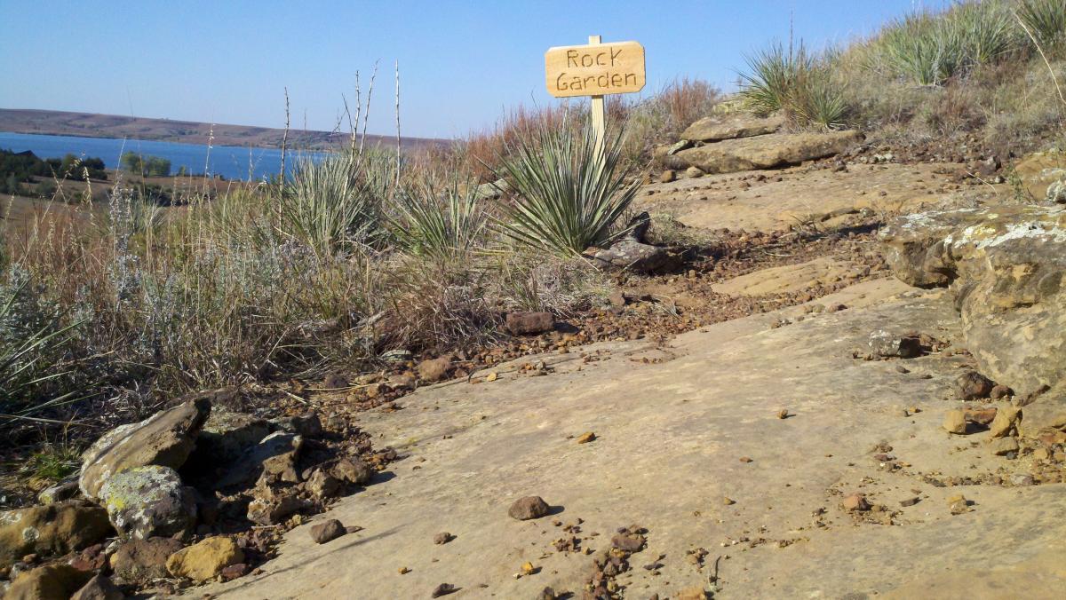 A rocky pathway leading to a scenic view of a lake, with a wooden sign marking the area as "Rock Garden." Surrounding vegetation includes grasses and scattered rocks on a clear blue sky day. Switchgrass mountain bike trail.