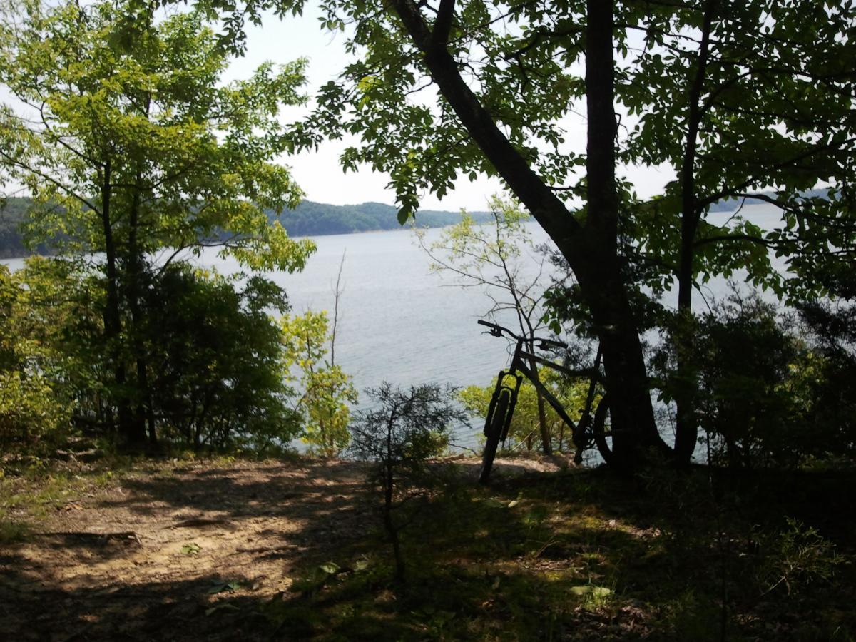 A peaceful lakeside scene framed by greenery, featuring a bike leaning against a tree. The calm water reflects the surrounding hills, creating a tranquil atmosphere in nature. Green River mountain bike trail.