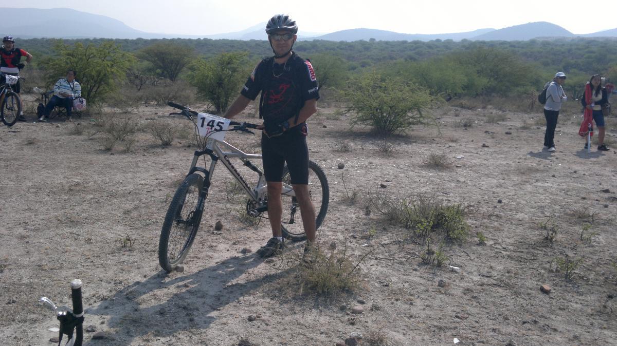 A mountain biker standing beside his bicycle in a barren landscape. He is wearing a helmet and athletic clothing with a number pinned to his shirt. In the background, several people are visible, and there are sparse shrubs and hills in the distance, suggesting an outdoor cycling event. Cerro Del Pitol mountain bike trail.