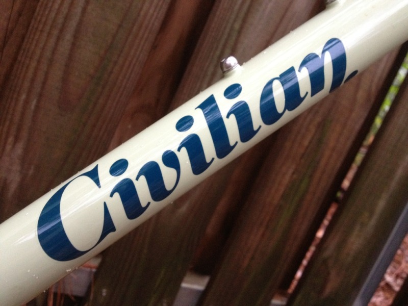 Close-up of a bicycle frame featuring the word "Civilian" in bold, navy blue lettering on a light-colored background, with wooden fence slats in the background.