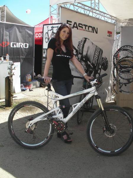 Airborne Zeppelin Elite: A woman poses next to a white mountain bike at an outdoor event, wearing a black shirt and dark jeans. She stands in front of a backdrop featuring bike accessories and logos, with various bicycle parts displayed in the vicinity. The setting appears to be a biking festival or exhibition.