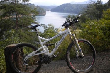Airborne Zeppelin Elite: A white mountain bike resting on a log, overlooking a scenic lake surrounded by lush greenery and hills in the background. The calm water reflects the landscape, creating a tranquil outdoor setting.
