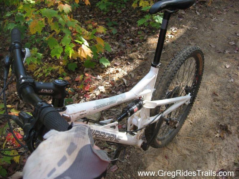 Airborne Zeppelin Elite: A close-up view of a white mountain bike positioned on a dirt trail surrounded by colorful autumn foliage. The handlebars and part of the frame are visible, showcasing the bike's design and components, including the seat and rear suspension. The ground is covered with fallen leaves, indicating a vibrant natural setting.