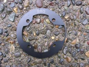 BBG Bashguard: A circular black metal piece with cutouts and holes, resting on a textured, pebbled surface.