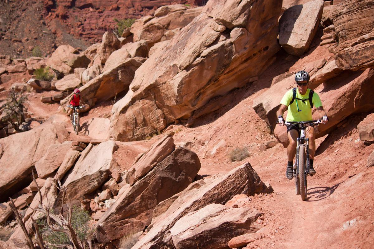 Two mountain bikers navigate a rocky trail in a desert landscape. One rider, dressed in a red jersey, is seen in the background, while the other, wearing a bright yellow shirt and a helmet, is riding on a narrow path in the foreground. Large boulders and red soil surround them under a clear blue sky. Moab Brand Trails mountain bike trail.