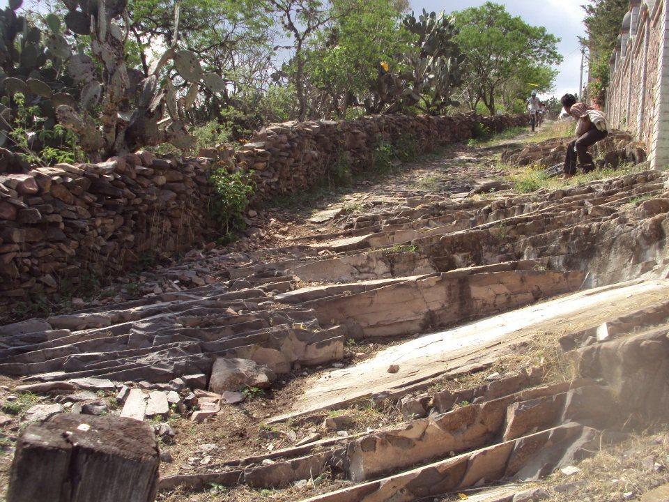 A rocky path lined with stone walls, featuring uneven, tiered terrain and sparse vegetation. Two individuals are visible in the background, one bending down and the other walking along the path, surrounded by cacti and trees under a partly cloudy sky. Paso De Mata, Sjr mountain bike trail.