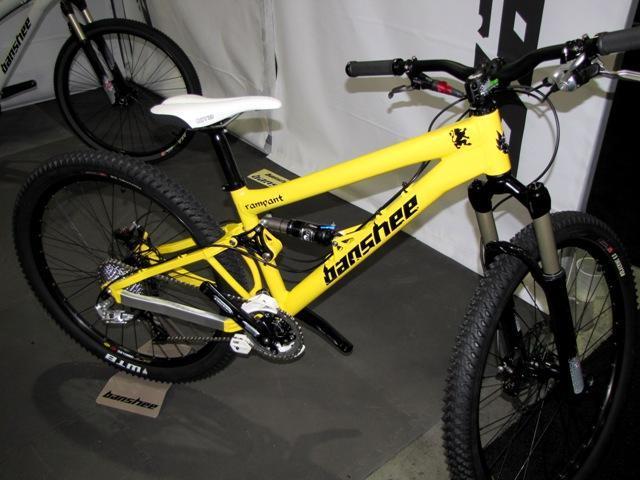Banshee Rampant: A bright yellow mountain bike with "Banshee" branding displayed in an exhibition area. The bike features a dual suspension system, knobby tires for off-road traction, and a sleek design. A white saddle is visible, along with various gears and components typical of high-performance mountain bikes.
