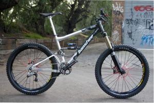 Banshee Rune: A silver and black mountain bike with oversized tires, a front suspension fork, and a sturdy frame, positioned on a paved surface with graffiti-covered walls and trees in the background.