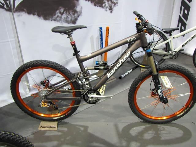 Banshee Rune: A high-performance mountain bike featuring a sleek gray frame and vibrant orange accents. It has thick, knobby tires designed for rugged terrain, a sturdy dual-suspension system, and high-quality components, showcasing its advanced engineering. The bike is displayed on a neutral background, emphasizing its design.