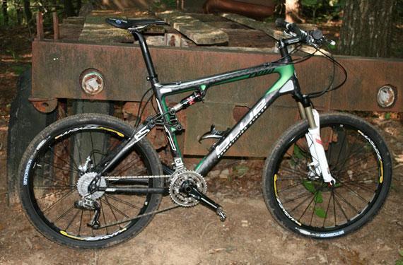 Blue Ryno Carbon: A black and green mountain bike resting on the ground, with a rugged, rusty machine in the background. The bike features thick tires, a lightweight frame, and a front suspension fork, set in a wooded area with natural vegetation.