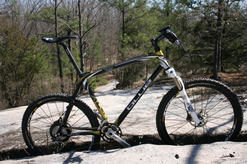 Blue XC Carbon: A mountain bike with a black and yellow frame is positioned on a rocky surface, surrounded by trees. The bike features thick tires suitable for off-road terrain and is set in a natural outdoor environment.
