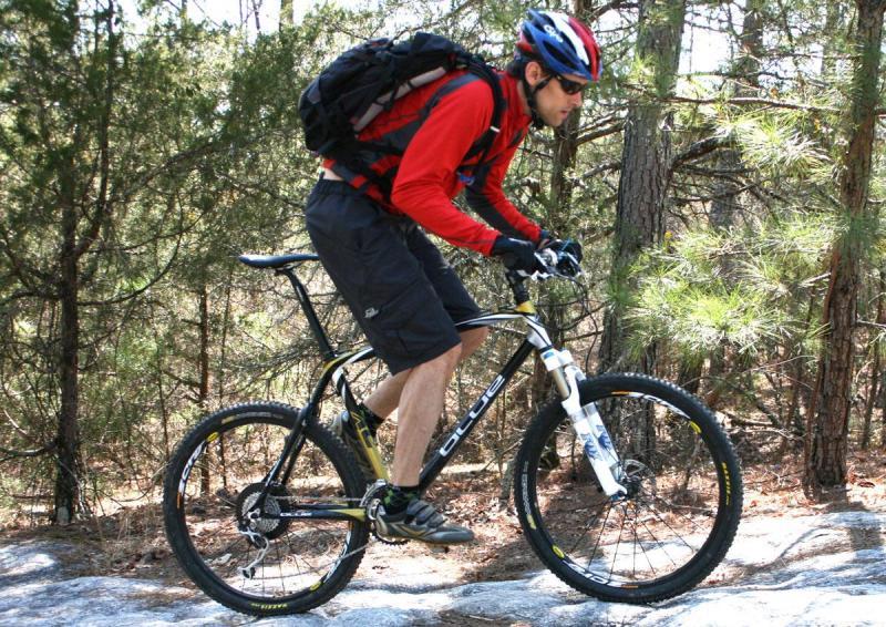 Blue XC Carbon: A man in a red long-sleeve shirt and black shorts rides a mountain bike on rocky terrain, surrounded by trees. He wears a helmet and sunglasses, and has a backpack secured on his back.