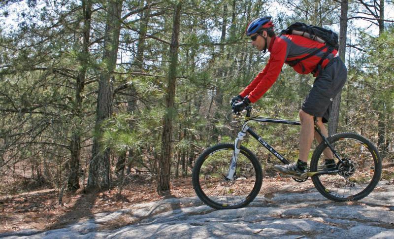 Blue XC Carbon: A mountain biker in a red shirt and gray shorts rides over rocky terrain in a forested area, surrounded by pine trees. The biker wears a helmet and has a backpack, focusing on navigating the natural landscape.