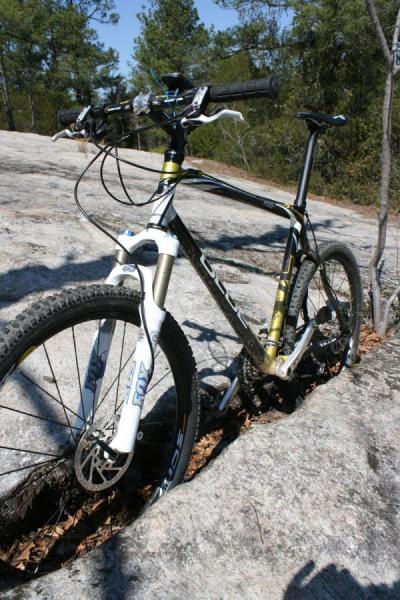 Blue XC Carbon: A black and white mountain bike positioned on a rocky surface, surrounded by trees. The bike features a sturdy frame, thick tires, and front suspension, indicating it's designed for off-road trails. Sunlight highlights the bike's details against the natural backdrop.