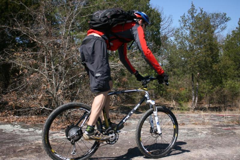 Blue XC Carbon: A cyclist in a red and black outfit rides a mountain bike on rocky terrain, surrounded by trees and brush. The cyclist is wearing a helmet and a backpack, leaning forward as they navigate the landscape.