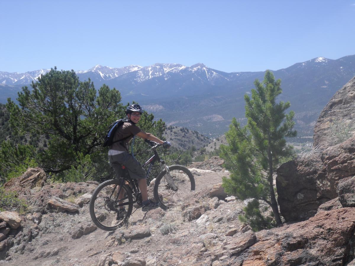 A person riding a mountain bike along a rocky trail, with mountains and trees in the background. The scene is set under a clear blue sky, suggesting a bright and sunny day. The rider is smiling and wearing a helmet, enjoying the outdoor adventure. Arkansas Hills mountain bike trail.