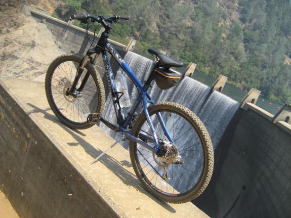 Cannondale F5: A blue mountain bike positioned on a concrete ledge near a dam, with a waterfall cascading in the background and lush greenery visible on the opposite bank. The bike has a water bottle attached to its frame and is facing towards the dam structure.