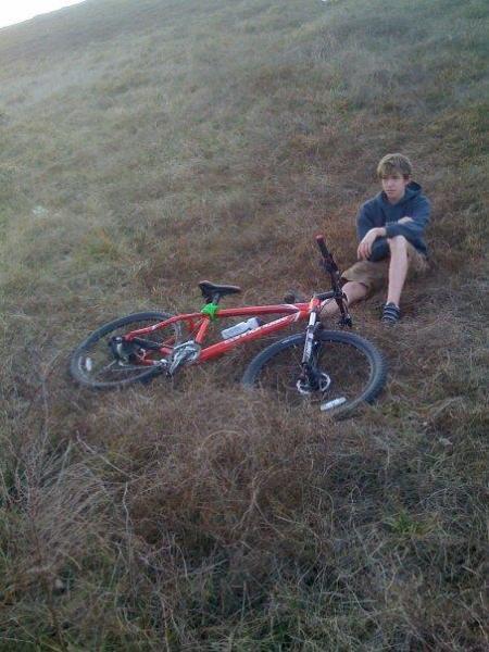 Cannondale F5: A boy sitting on the ground beside a red mountain bike, which is lying on its side in tall grass. The scene is set on a gentle slope under natural lighting. The boy appears to be relaxed while wearing shorts and a hoodie.