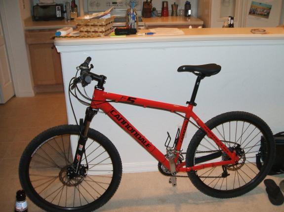 Cannondale F5: A red Cannondale mountain bike with a front suspension and thick tires, leaning against a wall in a room. In the background, there is a kitchen counter with items like a carton of eggs and various containers visible.