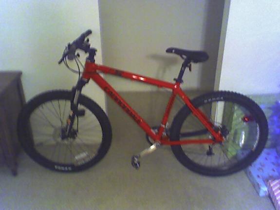 Cannondale F5: A red mountain bike with thick tires, standing on a light-colored carpet near a wall. The bike features a sturdy frame, handlebars, and disc brakes, with a small wooden side table visible in the background.
